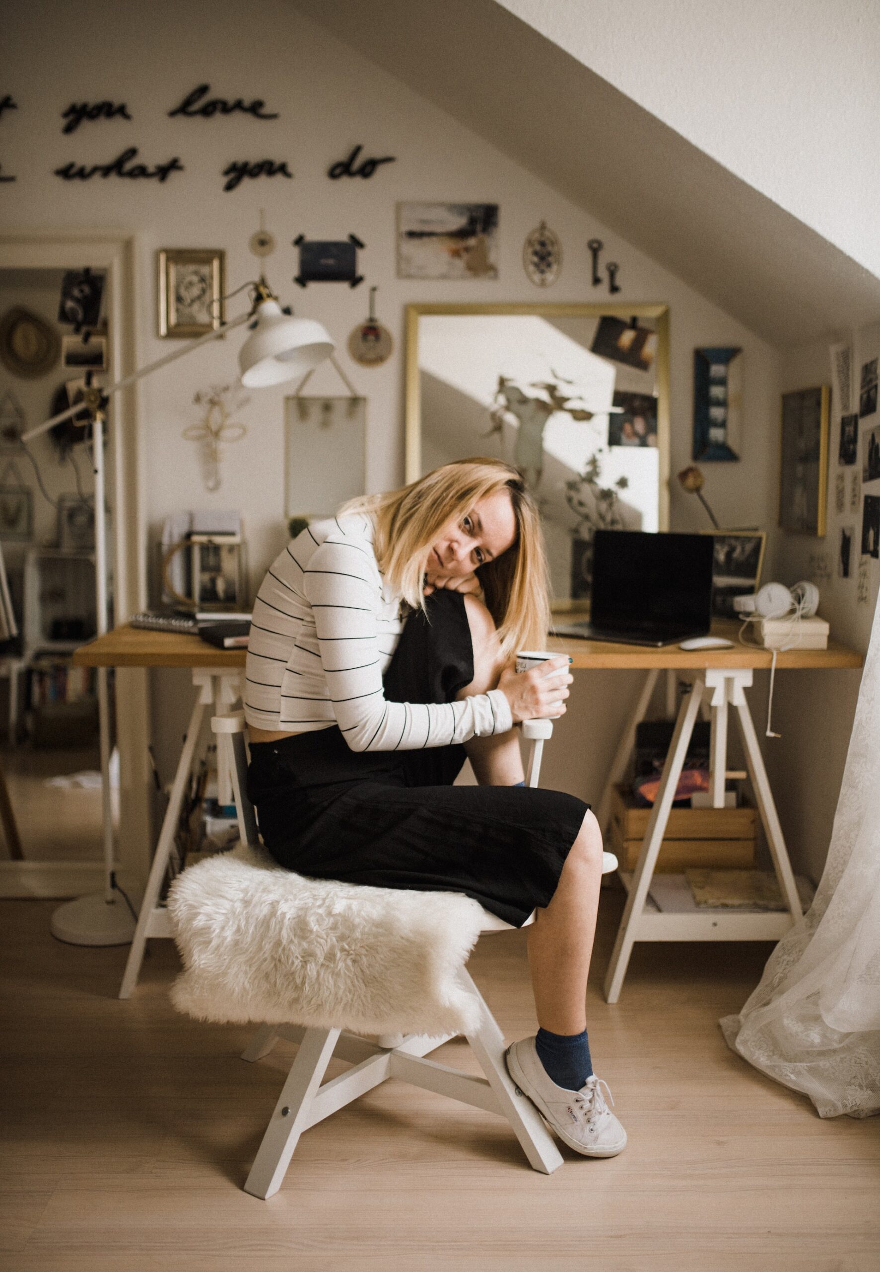 femme assise sur un tabouret dans un atelier en soupente, avec la tête appuyée sur un genou remonté et une tasse de café à la main